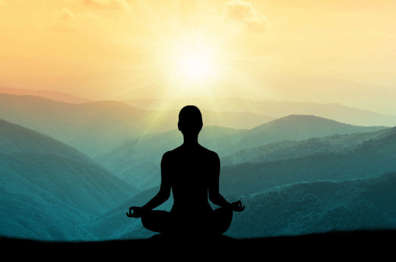 silhouette of person in lotus position looking out onto view of sun and mountains
