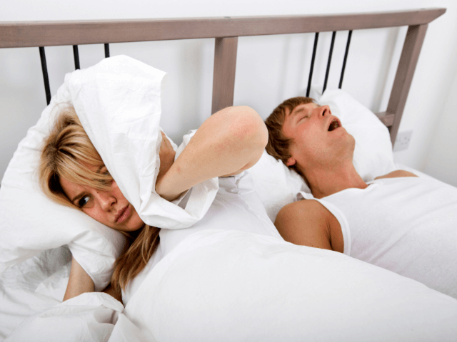 man snoring in bed while woman beside him covers ears with pillow